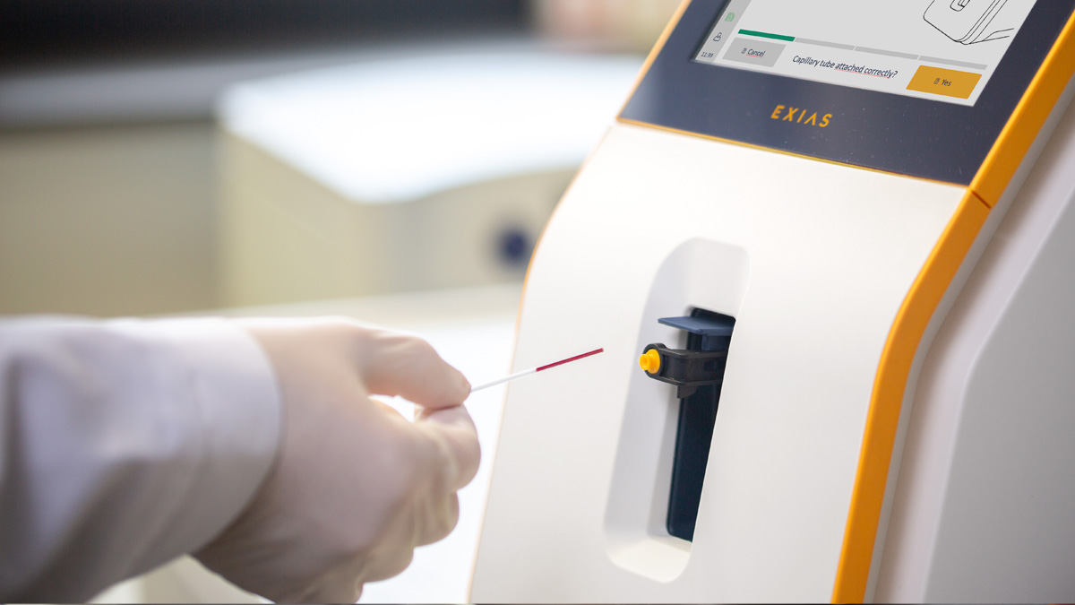 Hand i handskarna som sätter in ett blodprovrör i en medicinsk analysator med en skärm.