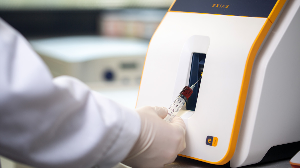 Hand med handskar för in en blodprovsspruta i en medicinsk analysator märkt "EXIAS" för testning.