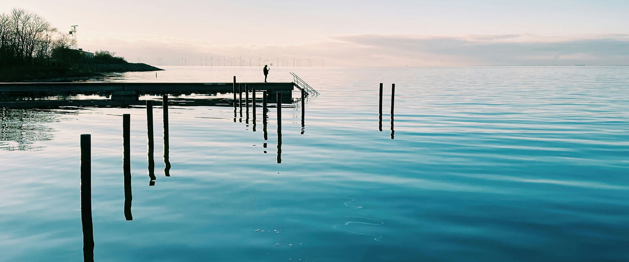 Brygga vid lugnt hav, person står i siluett, på morgonen med vindkraftverk i bakgrunden.