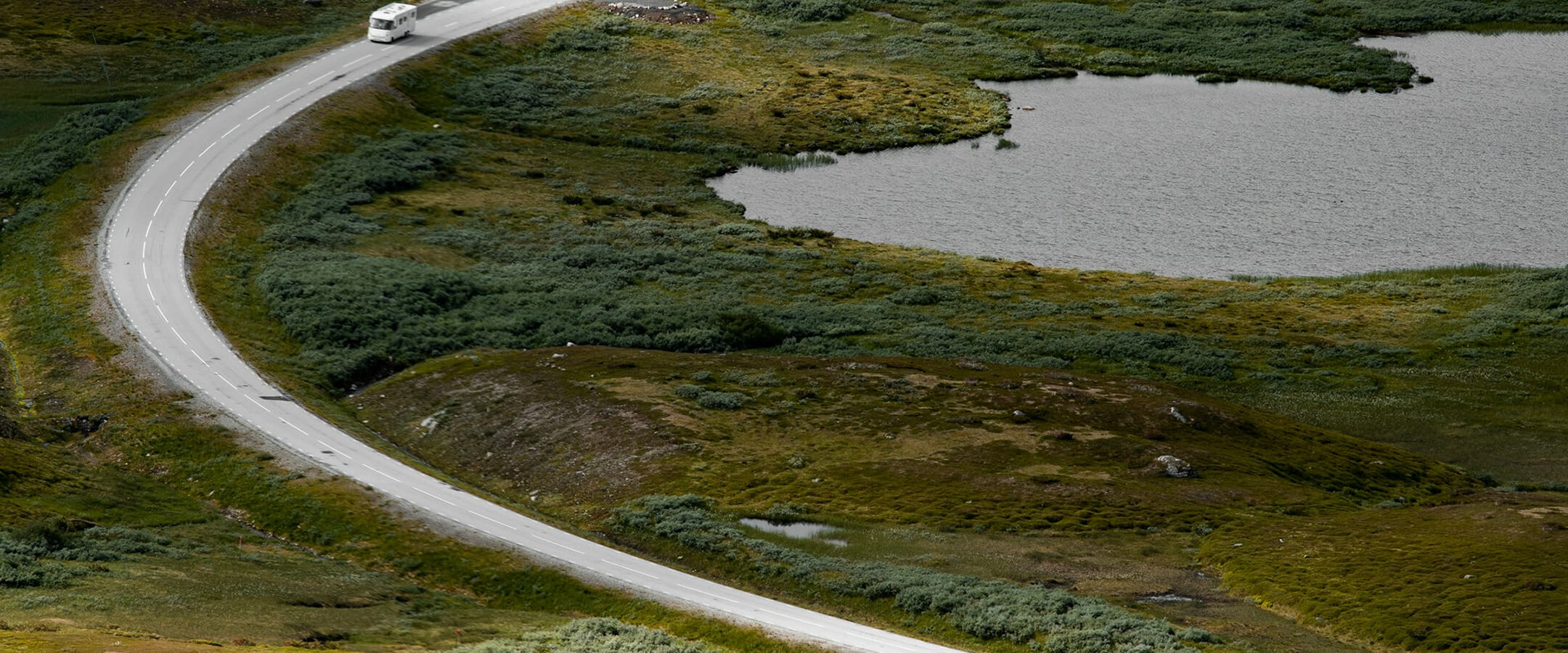 En vit husbil kör på en slingrande väg bredvid en sjö och grön vegetation.