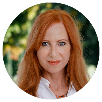 Kvinna med långt rött hår, vit skjorta och halsband, utomhus med suddig grön bakgrund.