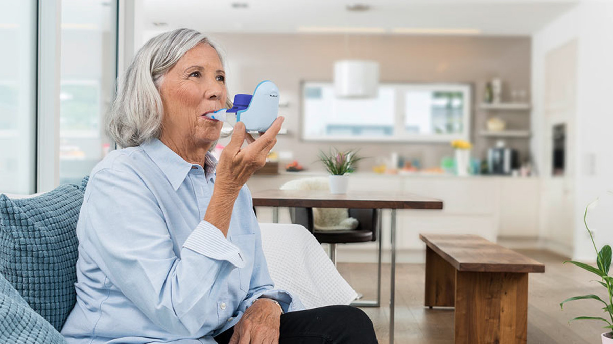Äldre kvinna som sitter på en soffa och använder en blå andningsinhalator i ett ljust modernt kök.