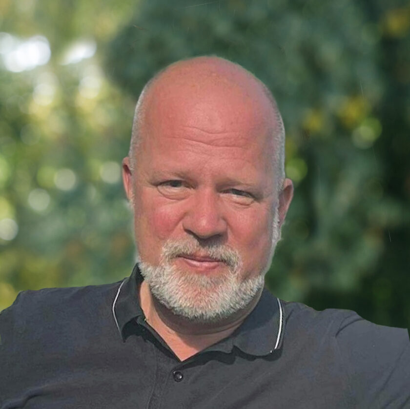 Skallig man med grått skägg i svart pikétröja, suddig grön bakgrund utomhus.