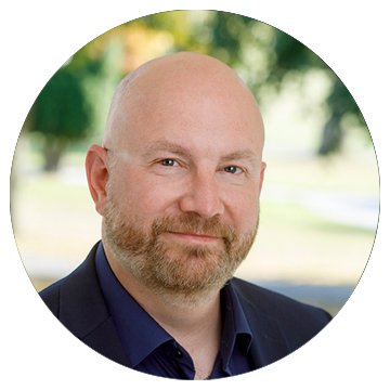 Skallig man med skägg, blå skjorta och kavaj, utomhus med suddig grön bakgrund.