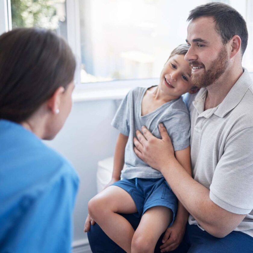 Doktor som undersöker en ung pojke som sitter i sin fars knä på ett sjukhus