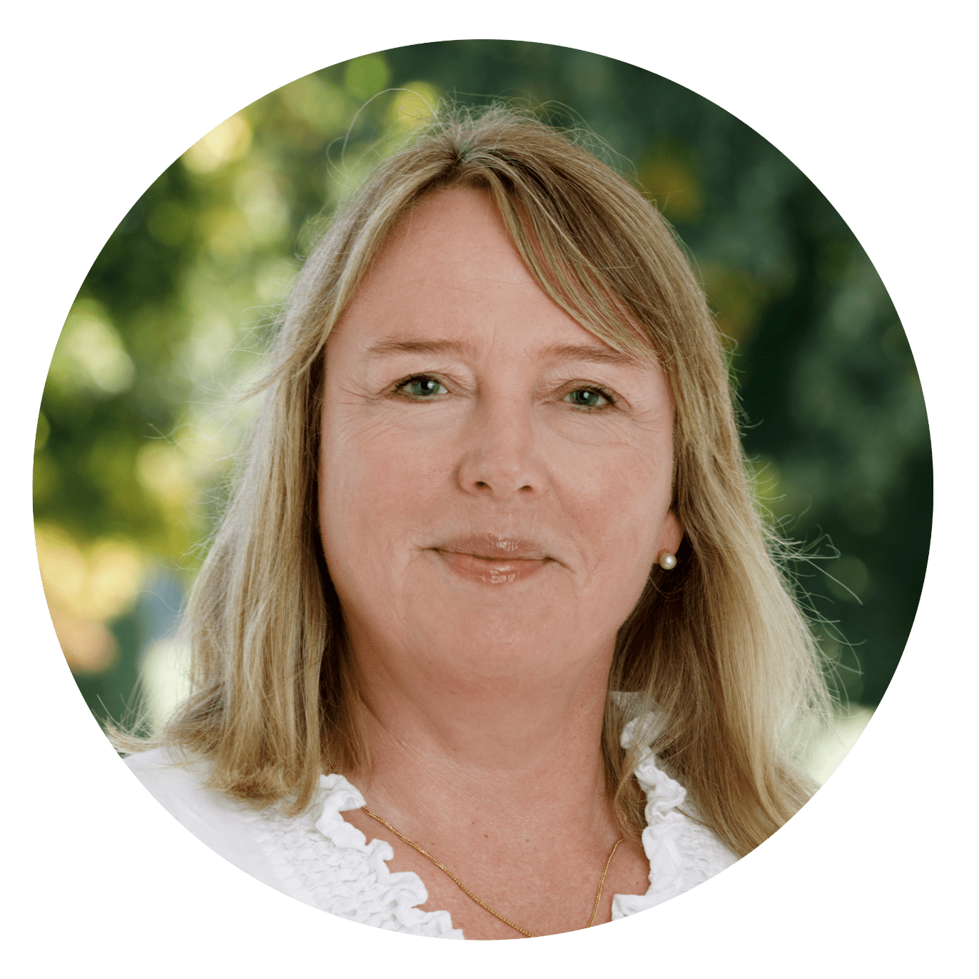 Kvinna med blont hår och vit blus, leende utomhus med suddig grön bakgrund.