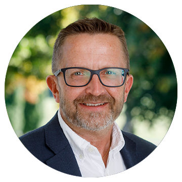 Man med glasögon och skägg ler mot kameran, bakgrund med gröna träd och solljus.