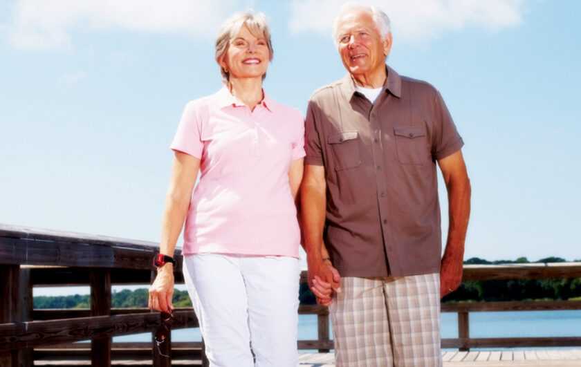 Ett äldre par håller hand och går på en brygga utomhus en solig dag.