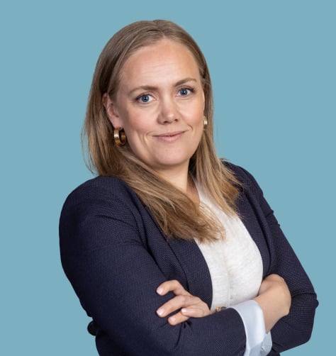 Kvinna med långt blondt hår i en marinblå kavaj och vit skjorta, armarna i kors, mot en blå bakgrund.