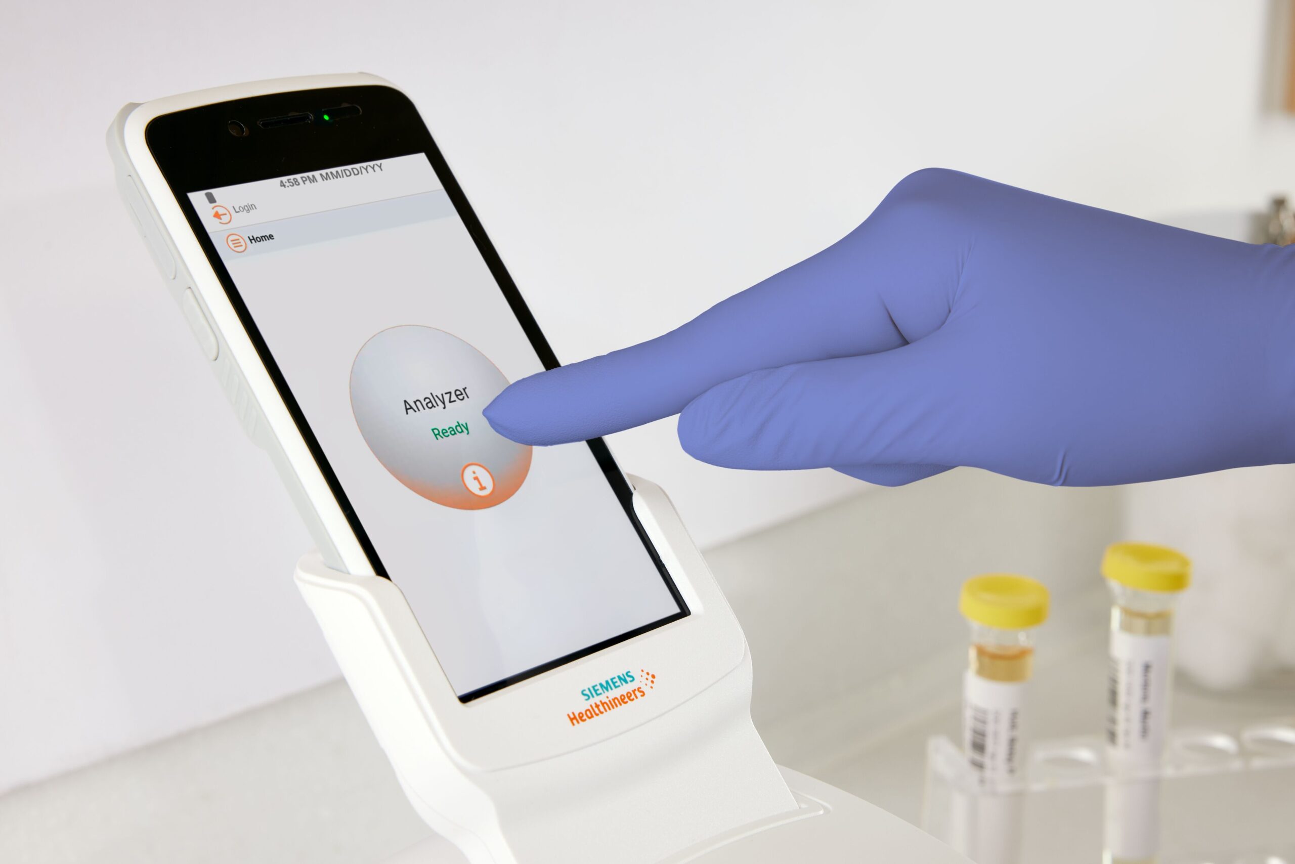 Handske försedd hand rör vid en Siemens Healthineers analysators pekskärm, med provrör i bakgrunden.
