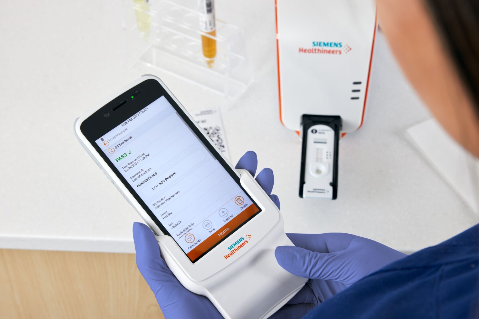Handskar klädda händer håller en Siemens Healthineers diagnostisk enhet som visar ett testresultat i en medicinsk laboratoriesituation.