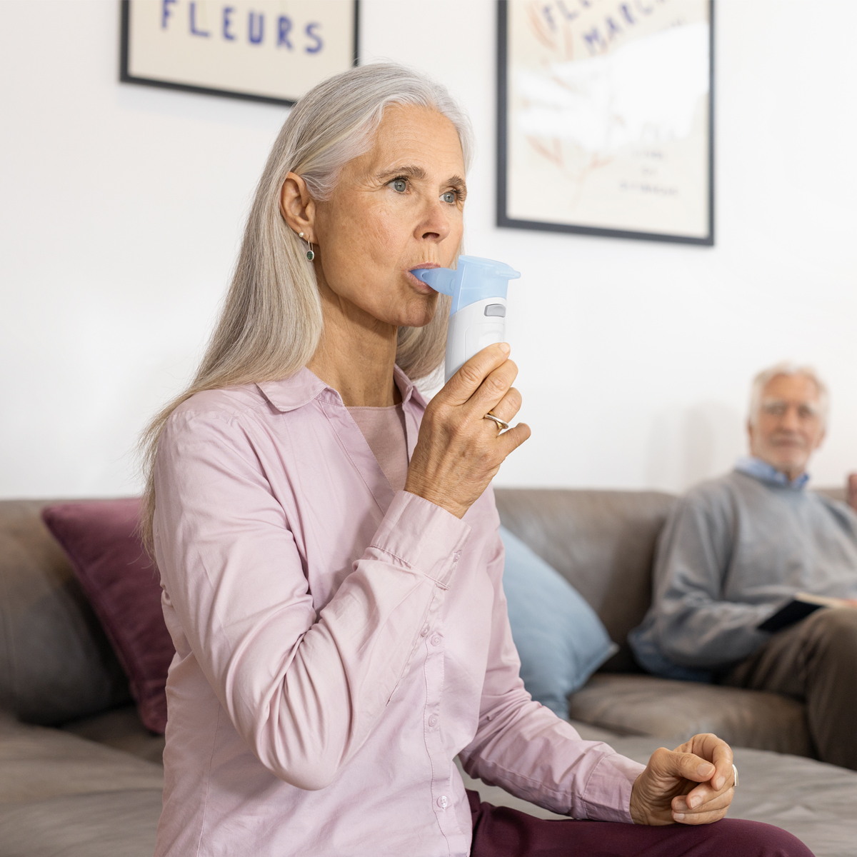 Äldre kvinna som använder en inhalator eller nebulisator medan hon sitter i en soffa, med en äldre man i bakgrunden.