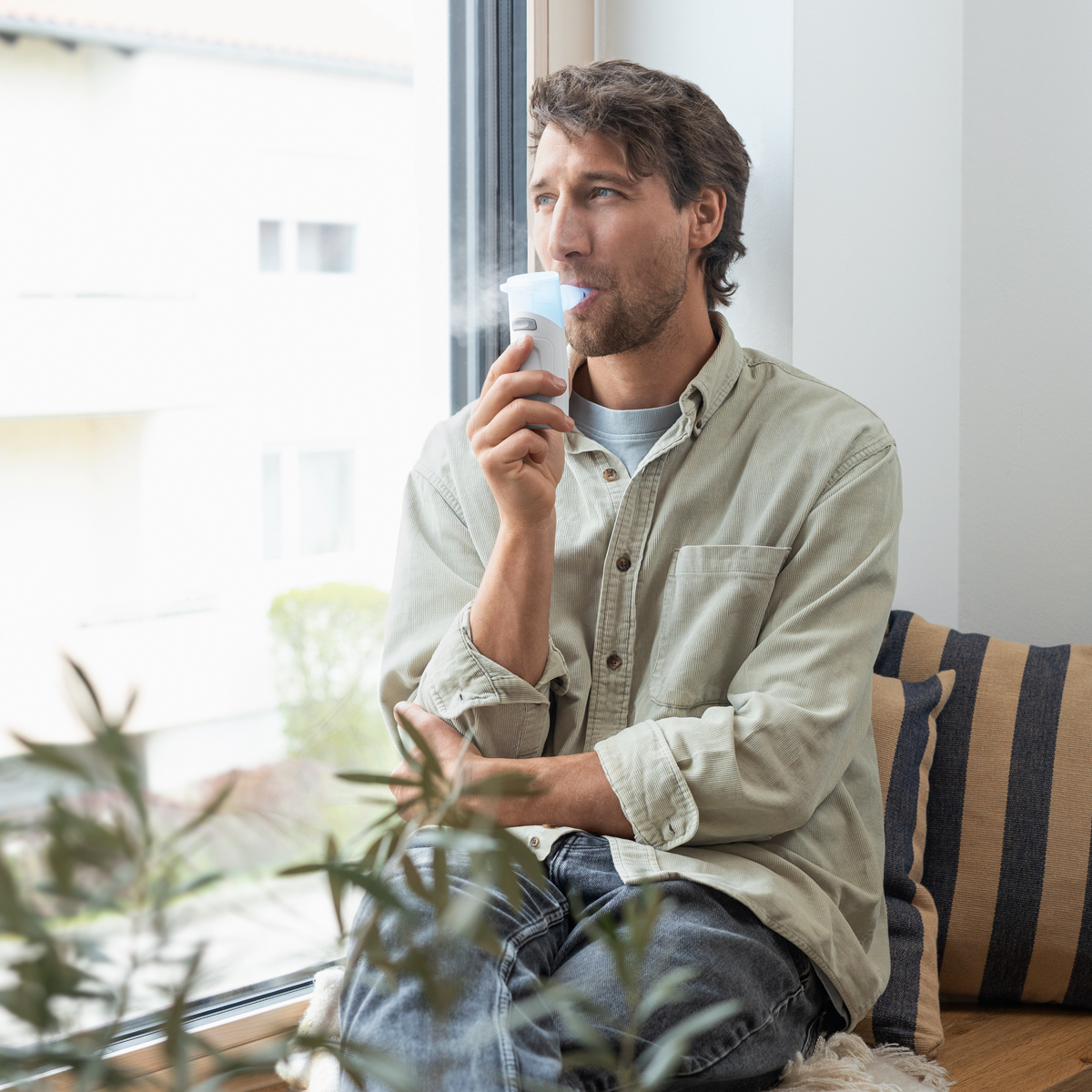 Man sitter vid ett fönster och använder en inhalator, bär en ljus skjorta, med växter i förgrunden.